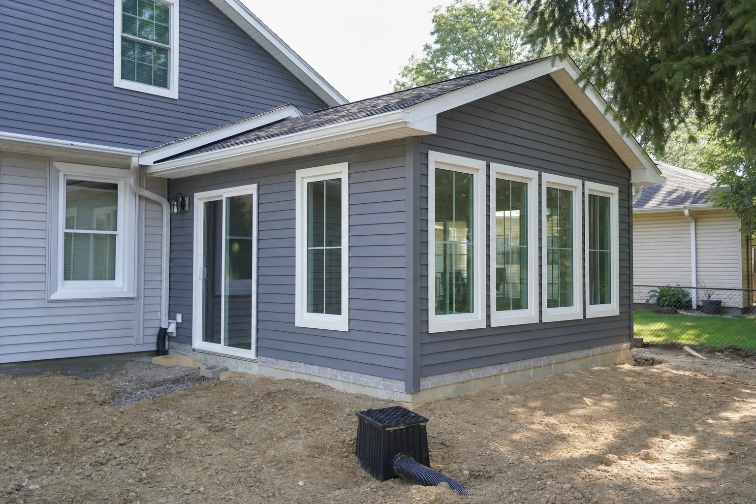 Newly constructed gray vinyl siding home addition with large white-trimmed windows on bare dirt lot during landscaping phase.